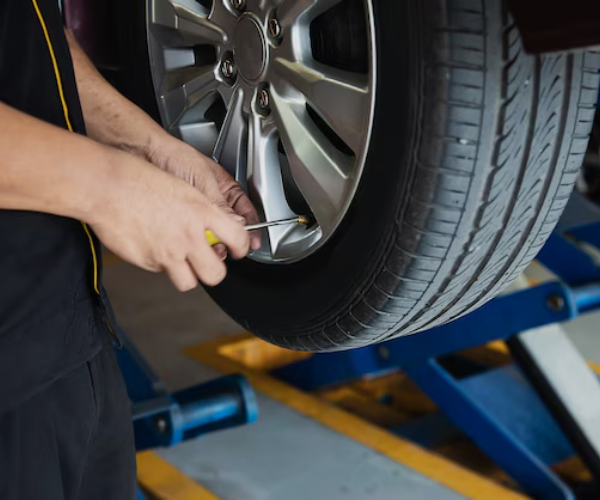 A mechanic working on a car's wheel, possibly checking the tire pressure or adjusting the valve stem. The car is lifted on a hydraulic platform in an automotive repair shop.