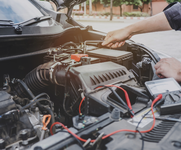 Two hands work on a car engine, using a diagnostic tool with red and black cables, under an open hood in a sunny outdoor setting.