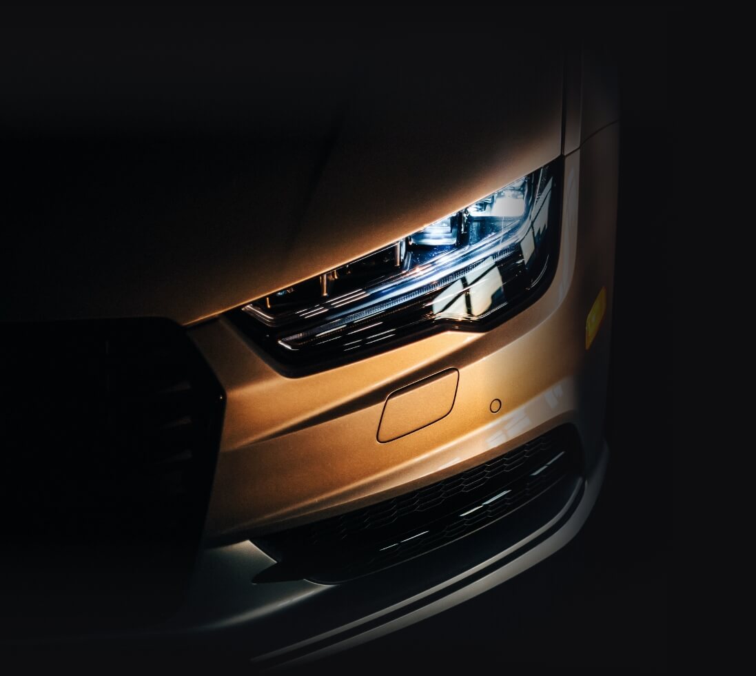 Close-up of a sleek bronze car's front with glowing headlights in a dimly lit setting.