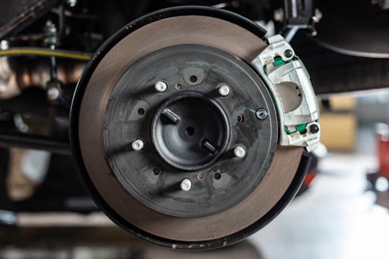 A close-up view of a car brake disc and caliper under the vehicle, showing the brake system components in a garage setting with tools and parts in the background.