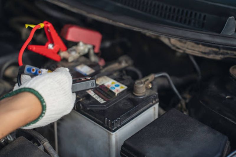  A gloved hand connecting red and black jumper cables to a car battery under the hood, showing a close-up of the battery and engine compartment during a jump-start process.