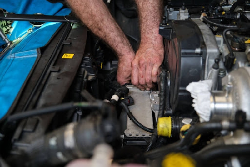 A pair of hands working on a car engine under the hood, adjusting or repairing electrical components and wires in a garage with tools and parts visible.