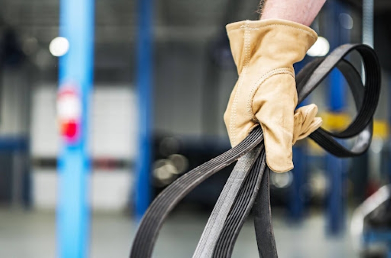 A gloved hand holding a car belt, likely from the engine, in a garage with blue pillars and blurred tools and vehicles in the background.