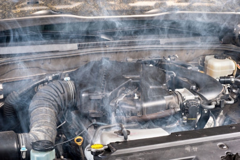 A car engine under the hood releasing steam, indicating an overheating issue, with visible components like hoses and the coolant cap in a garage setting.
