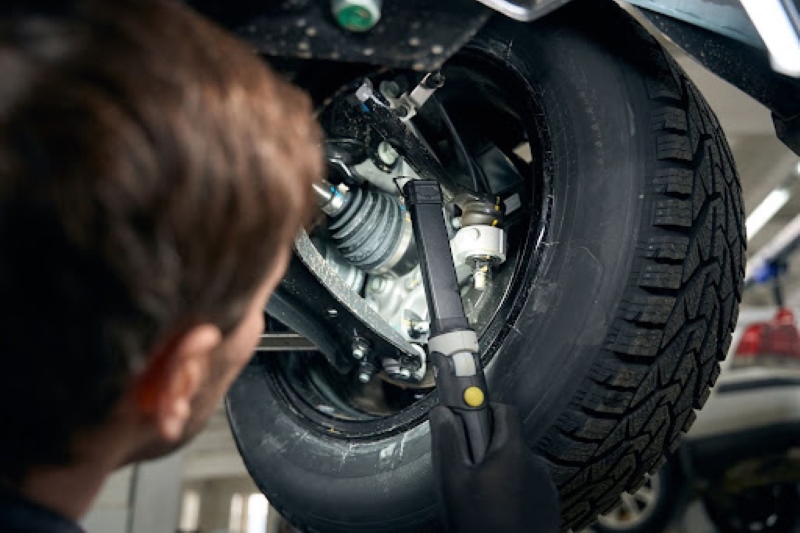 A person with gloved hands using a tool to work on a car’s suspension system, with the vehicle lifted and a tire visible in a garage environment.