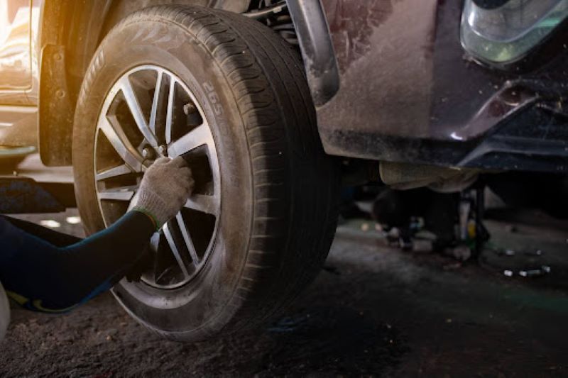 A gloved hand working on a car tire, adjusting or removing it from a vehicle lifted in a garage, with sunlight streaming in and tools visible in the background.