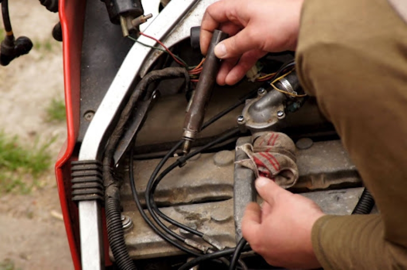 A hand holding a spark plug while working on a motorcycle engine, with wires and components visible under the hood in an outdoor setting.
