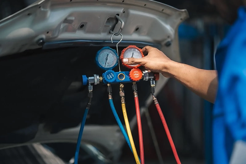 Mechanic adjusting manifold gauge to check car air conditioning system under open hood in auto repair shop.