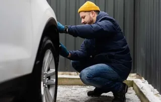 A man in a yellow beanie and blue jacket performing a manual tyre tread and pressure check on a white car.